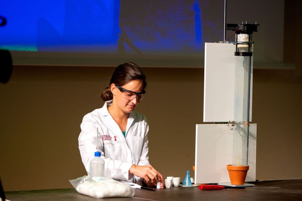 Eine Frau mit zusammengebundenen Haaren trägt einen weißen Laborkittel und eine Schutzbrille. Sie sitzt an einem Tisch und hantiert mit kleinen Laborutensilien, darunter ein Becherglas und ein weißer Behälter. Auf dem Tisch liegen weitere Laborgegenstände, darunter ein Trichter, ein Tongefäß und eine rote Pinzette. Im Hintergrund steht ein hohes, weißes Versuchsaufbau-Gerät mit Messskala und einem kleinen Behälter an der Spitze. Die Szene ist in einem Labor oder bei einer wissenschaftlichen Vorführung aufgenommen.