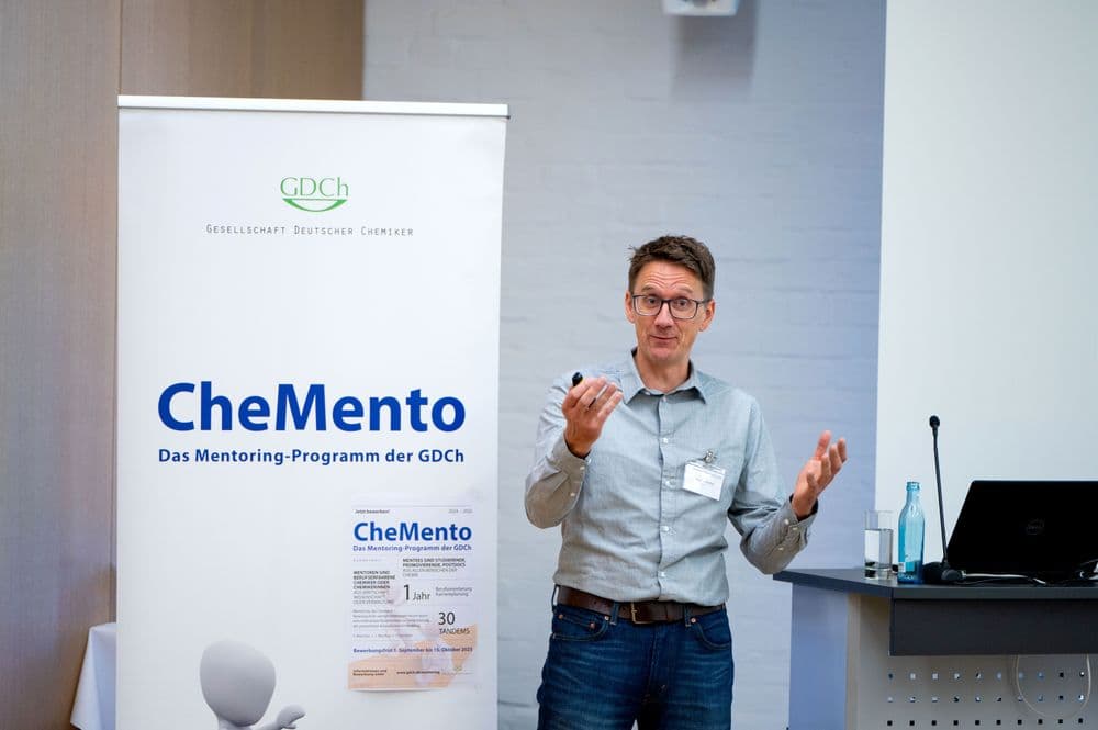 Ein Mann mit Brille, grauem Haar und Namensschild steht vor einem Roll-up-Banner und hält einen Vortrag. Er trägt ein hellblaues Hemd und eine dunkle Hose. In der rechten Hand hält er einen Präsentationsklicker, die linke Hand ist leicht erhoben. Das Banner zeigt das Logo der Gesellschaft Deutscher Chemiker und den Schriftzug 'CheMento Das Mentoring-Programm der GDCh'. Im Hintergrund stehen ein Laptop, eine Glasflasche und ein Glas auf einem Tisch.