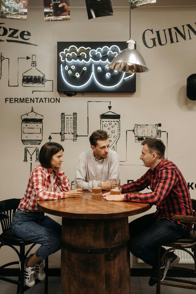 Drei Personen sitzen an einem runden Holztisch in einem Café oder einer Bar. Zwei tragen karierte Hemden, eine Person ein helles Hemd. Auf dem Tisch stehen Gläser mit Getränken. Die Wand im Hintergrund ist mit technischen Zeichnungen von Brauanlagen, englischen Begriffen wie 'FERMENTATION' und 'GUINNESS' sowie einer leuchtenden Wandinstallation dekoriert. Eine silberne Hängelampe beleuchtet die Szene.
