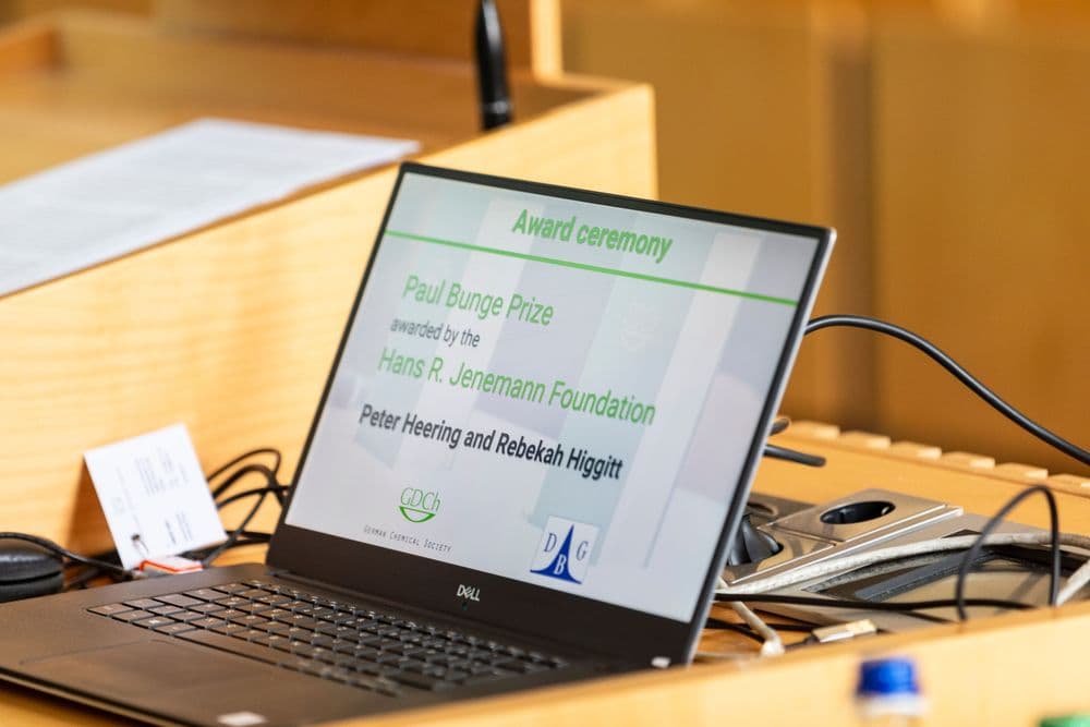 Ein aufgeklappter Laptop auf einem Holzpult zeigt eine Präsentationsfolie mit der Überschrift 'Award ceremony'. Darauf steht: 'Paul Bunge Prize awarded by the Hans R. Jenemann Foundation, Peter Heering and Rebekah Higgitt'. Unten auf der Folie sind die Logos der GDCh (German Chemical Society) und der DBG zu sehen. Im Hintergrund liegen Kabel und ein Blatt Papier auf dem Pult.
