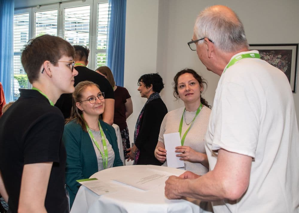 Im Vordergrund stehen drei Frauen und ein Mann um einen Stehtisch, alle tragen grüne Namensschilder um den Hals. Die Frau links hat kurze Haare und eine Brille, die Frau daneben trägt ein türkisfarbenes Jackett und ebenfalls eine Brille. Die dritte Frau hat dunkle, lockige Haare und hält ein Blatt Papier in der Hand. Der Mann rechts ist grauhaarig, trägt eine Brille und ein helles T-Shirt. Im Hintergrund unterhalten sich weitere Personen in einem hellen Raum mit großen Fenstern und blauen Vorhängen. Laut Kontext handelt es sich bei den Personen von links nach rechts um Bärbel Arling, Barbara Pohl und Peter Staniek.