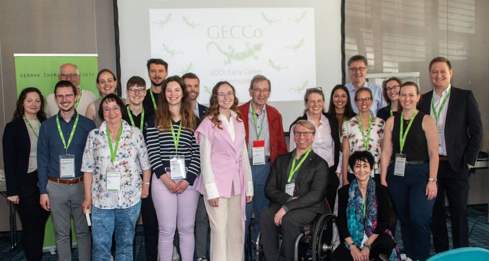 Eine Gruppe von Menschen steht in einem Konferenzraum vor einer Leinwand mit dem projizierten Schriftzug 'GECCo' und einem grünen Gecko-Logo. Die Personen tragen Namensschilder und grüne Lanyards. Im Vordergrund stehen drei Personen, von links nach rechts: Bärbel Arling, Barbara Pohl und Peter Staniek. Peter Staniek sitzt im Rollstuhl. Die Gruppe wirkt freundlich und professionell, einige Personen lächeln in die Kamera. Im Hintergrund sind Fenster mit Jalousien und ein grünes Roll-up mit dem Text 'German Chemical ...' sichtbar.