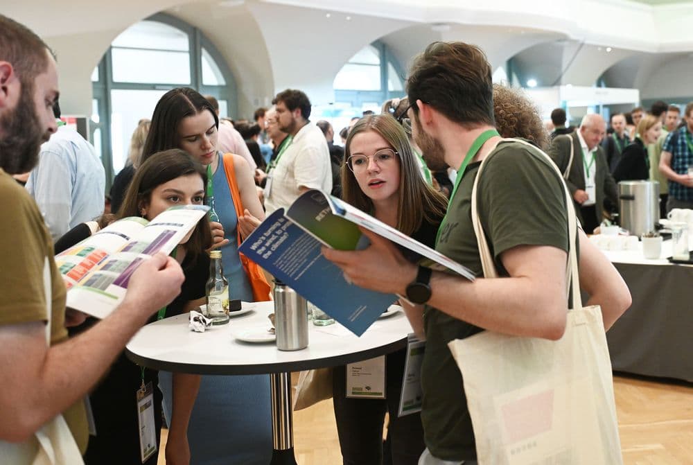 Menschen stehen um einen runden Stehtisch in einem hellen, modernen Raum. Einige blättern in Broschüren oder Magazinen, andere hören aufmerksam zu. Die Personen tragen Namensschilder und Umhängetaschen. Im Hintergrund sind weitere Menschen und Tische mit Getränken und Geschirr zu sehen. Die Atmosphäre wirkt geschäftig und kommunikativ.