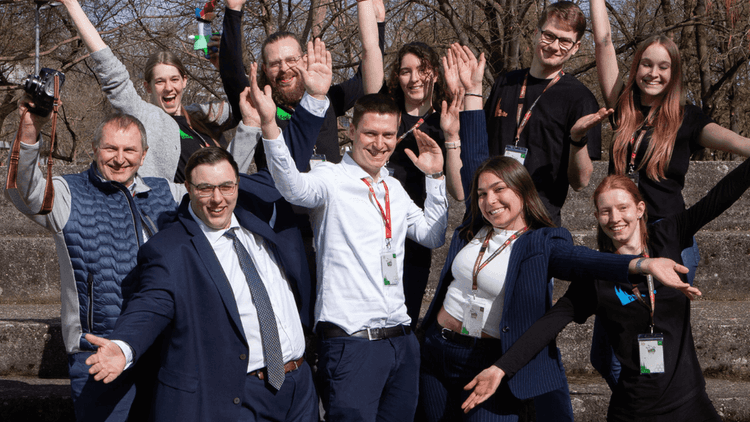 Eine Gruppe von Menschen steht auf steinernen Stufen im Freien. Alle tragen Namensschilder und heben die Arme, einige lächeln. Die Kleidung reicht von Anzügen bis zu legeren Outfits. Im Hintergrund sind Bäume ohne Laub und ein Gebäude mit Dach zu sehen. Das Bild vermittelt eine fröhliche, gemeinschaftliche Atmosphäre.