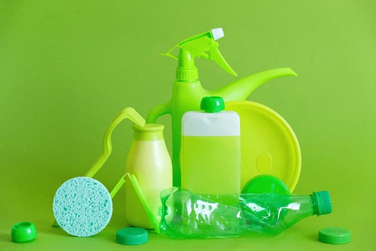 Verschiedene Haushaltsgegenstände aus Kunststoff stehen vor einem einfarbigen, hellgrünen Hintergrund. Zu sehen sind eine Gießkanne, eine Flasche mit Sprühkopf, eine Trinkflasche mit Henkel, ein Spülschwamm, eine flache Flasche mit weißem Verschluss, ein rundes Tablett und mehrere Kunststoffdeckel. Alle Gegenstände sind in unterschiedlichen Grüntönen gehalten.