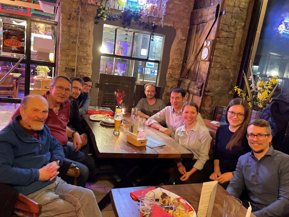 Eine Gruppe von Menschen sitzt um einen großen Holztisch in einem Restaurant oder einer Gaststätte. Die Szene zeigt den GDCh-VCW-Stammtisch. Auf dem Tisch stehen Getränke, darunter Bier und Wasser, sowie Teller mit Speisen und eine kleine Vase mit roten Blumen. Die Personen tragen überwiegend legere Kleidung und lächeln in Richtung Kamera. Im Hintergrund sind Steinwände, dekorative Pflanzen und eine beleuchtete Tür zu sehen. Die Atmosphäre wirkt entspannt und gesellig.
