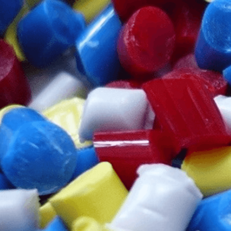 Bunte Kunststoffgranulate in quadratischer Form liegen dicht beieinander. Die Farben Blau, Rot, Gelb und Weiß sind deutlich zu erkennen. Die Oberfläche der Granulate wirkt glatt und glänzend.