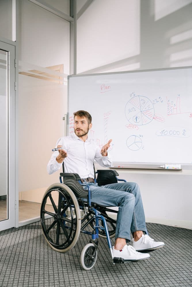 Ein Mann im Rollstuhl sitzt vor einem Whiteboard, das mit farbigen Diagrammen und Zahlen beschriftet ist. Er trägt ein weißes Hemd und helle Jeans, hält einen Marker in der Hand und gestikuliert mit beiden Armen. Der Raum wirkt modern und hell, mit Glaswänden und Teppichboden.