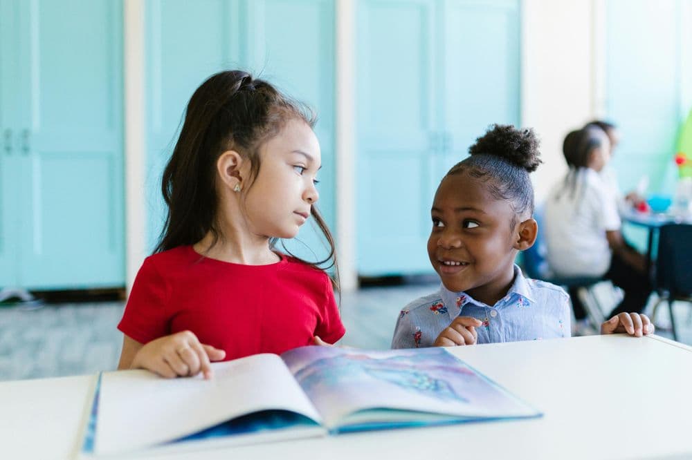 Zwei Kinder sitzen zusammen an einem Tisch und betrachten ein aufgeschlagenes Bilderbuch. Ein Mädchen mit langen dunklen Haaren trägt ein rotes T-Shirt, neben ihr sitzt ein Kind mit lockigen Haaren und einem gestreiften Hemd. Beide lächeln und scheinen sich über das Buch zu unterhalten. Im Hintergrund sind türkisfarbene Vorhänge und weitere Personen in einem Klassenraum zu sehen.