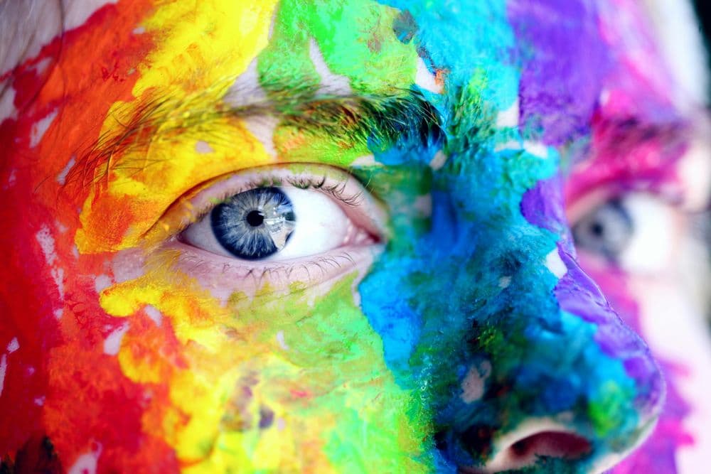 Nahaufnahme eines Gesichts mit intensiver, bunter Bemalung. Die Farben verlaufen in kräftigen Streifen und Flecken über die Haut und reichen von Gelb, Rot und Orange bis zu Blau, Grün und Violett. Das Auge in der Bildmitte ist klar erkennbar und wirkt durch die Bemalung besonders hervorgehoben. Die Struktur der Farbe ist stellenweise dick und unregelmäßig aufgetragen.